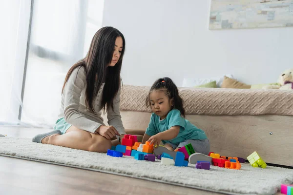 Chica asiática jugando bloques de construcción cerca de la madre en la alfombra en el suelo - foto de stock