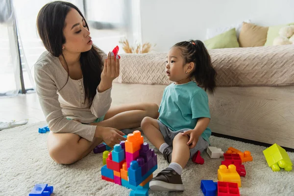 Asiática madre buscando en bloque de construcción cerca de hija en alfombra - foto de stock