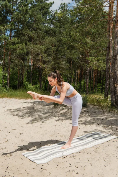 Cheerful and barefoot woman in sportswear stretching on yoga mat in forest — Stock Photo