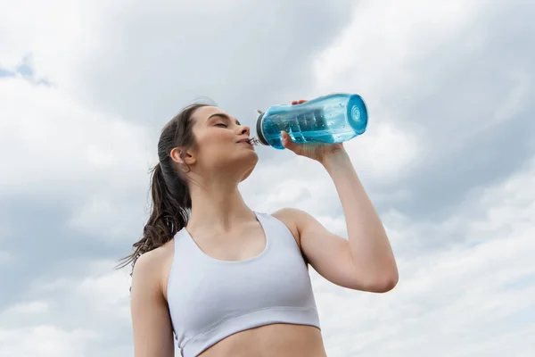 Low angle view of brunette woman in crop top drinking water against  sky with clouds — Stock Photo