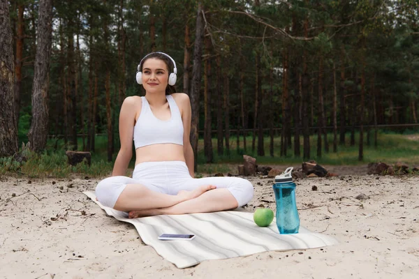 Mujer feliz en auriculares escuchando música mientras está sentada con las piernas cruzadas en la esterilla de yoga - foto de stock