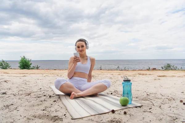 Smiling woman in wireless headphones listening music while using smartphone and sitting with crossed legs on yoga mat — Stock Photo