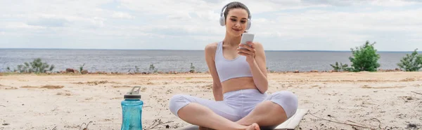 Donna sorridente in cuffia ascoltare musica durante l'utilizzo di smartphone e seduto con le gambe incrociate su tappetino yoga, banner — Foto stock