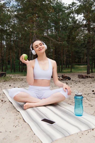 Щаслива жінка в навушниках слухає музику, тримаючи яблуко і сидячи з схрещеними ногами на килимку для йоги — стокове фото