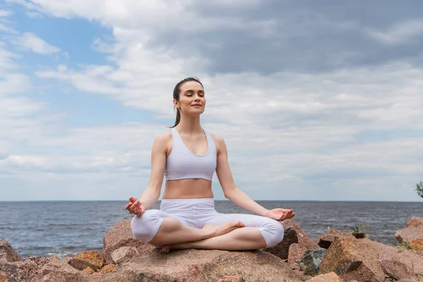 Cheerful woman in headphones and sportswear sitting in lotus pose while meditating near sea — Stock Photo