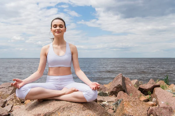 Pleased young woman in headphones and sportswear sitting in lotus pose while meditating near sea — Stock Photo