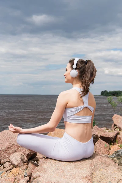 Happy woman in headphones and sportswear sitting in lotus pose while meditating near sea — Stock Photo