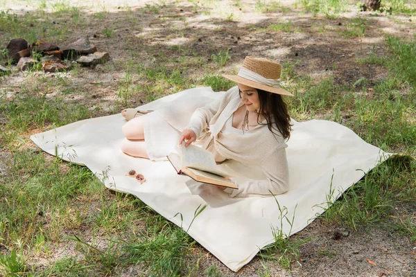 Mujer joven positiva en sombrero de paja libro de lectura en el bosque - foto de stock