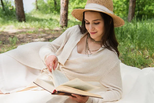 Jovem mulher positiva em chapéu de palha leitura romance na floresta — Fotografia de Stock