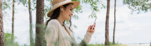 Positive young woman in straw hat and wireless earphones using smartphone in forest, banner — Stock Photo