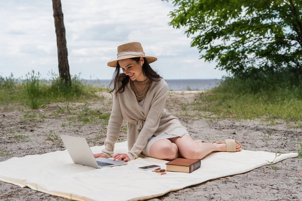 Mujer feliz en sombrero de paja y auriculares usando el ordenador portátil mientras está sentado en la manta de picnic cerca de teléfono inteligente, gafas de sol y libro - foto de stock