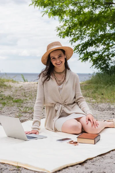 Mujer alegre en sombrero de paja y auriculares usando el ordenador portátil mientras está sentado en la manta de picnic cerca de teléfono inteligente, gafas de sol y libro - foto de stock