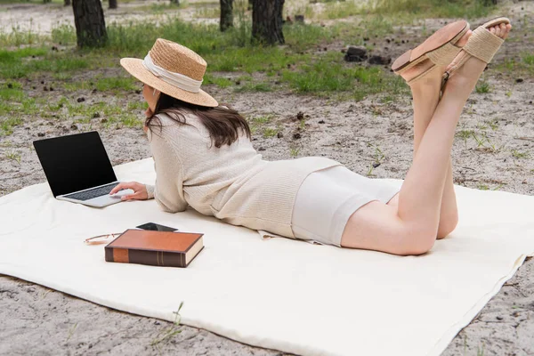 Woman in straw hat and earphones using laptop with blank screen while lying on picnic blanket near smartphone, sunglasses and book — Stock Photo