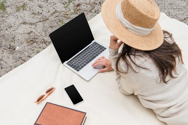 Vista de ángulo alto de la mujer en sombrero de paja usando el ordenador portátil con pantalla en blanco mientras está acostado en la manta de picnic cerca de teléfono inteligente, gafas de sol y libro - foto de stock