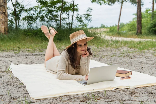 Улыбающаяся женщина в соломенной шляпе с помощью ноутбука, лежащего на одеяле для пикника рядом с солнцезащитными очками и книги — стоковое фото