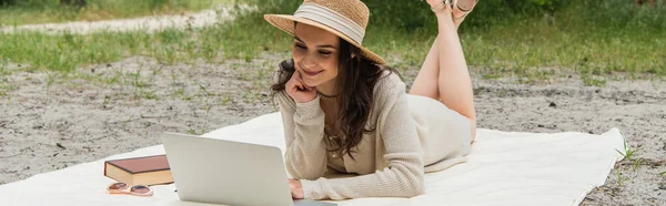 Усміхнена жінка в солом'яному капелюсі, використовуючи ноутбук, лежачи на ковдрі для пікніка біля сонцезахисних окулярів і книги, банер — стокове фото