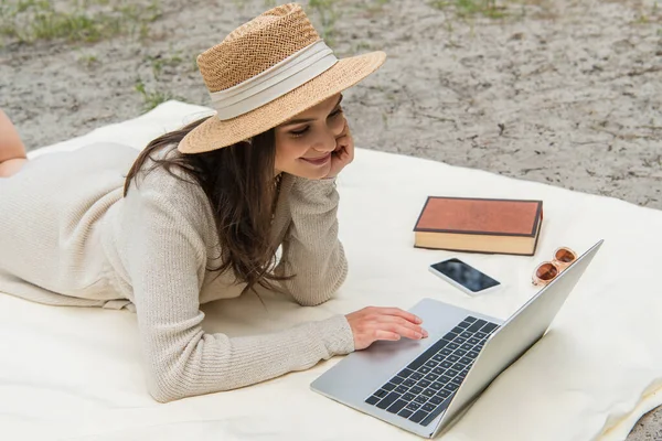 Alegre freelancer en sombrero de paja usando el ordenador portátil mientras está acostado en la manta de picnic cerca de smartphone, gafas de sol y libro - foto de stock