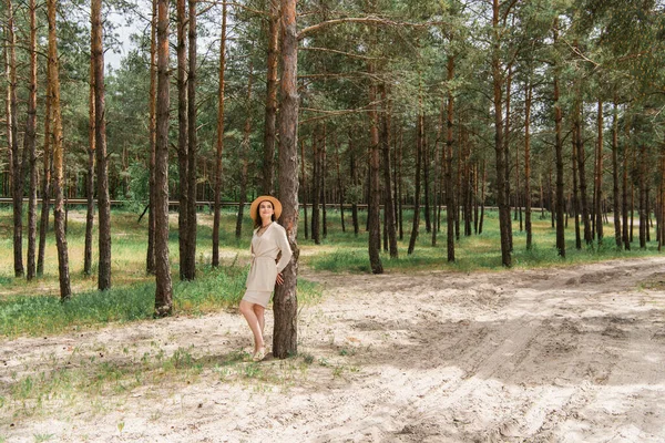 Full length of cheerful young woman in straw hat standing in woods — Stock Photo