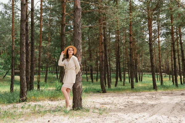 Longitud completa de alegre joven mujer ajustando sombrero de paja y de pie en el bosque - foto de stock