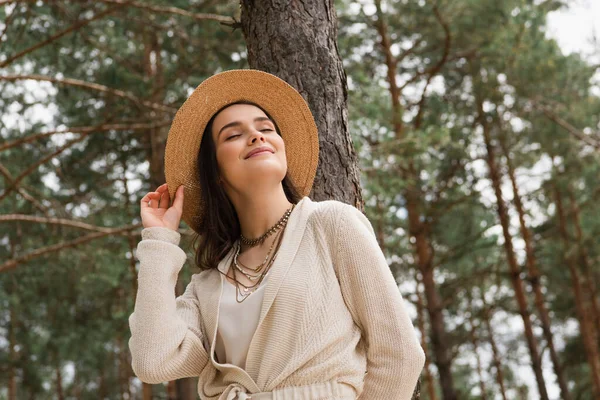 Cheerful young woman adjusting straw hat and smiling in woods — Stock Photo