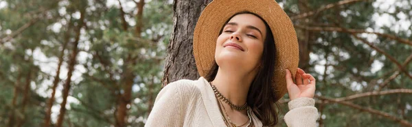 Весела молода жінка регулює солом'яний капелюх і посміхається в лісі, банер — стокове фото