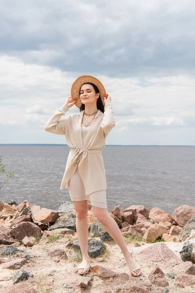 Pleine longueur de sourire jeune femme ajustant chapeau de paille et debout près de la mer — Photo de stock