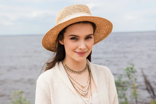 Heureux jeune femme en chapeau de soleil regardant la caméra près de la mer — Photo de stock
