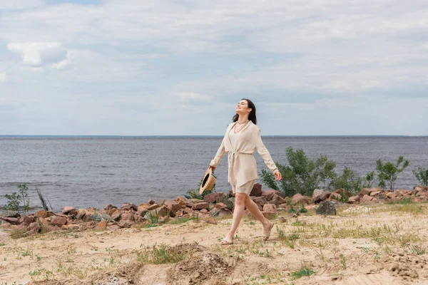 Longitud completa de sonriente mujer joven sosteniendo sombrero de paja y caminando cerca del mar - foto de stock