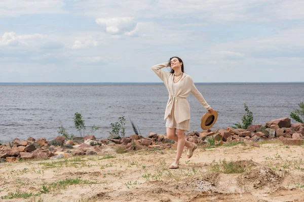 Volle Länge der glücklichen jungen Frau mit Sonnenhut und Fuß in der Nähe des Meeres — Stockfoto