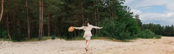 Вид сзади молодой женщины с протянутыми руками, держащей соломенную шляпу и идущей возле леса, баннер — стоковое фото