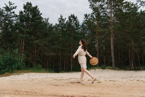 Pleine longueur de brune jeune femme en lunettes de soleil tenant chapeau de paille et marchant près de la forêt — Photo de stock