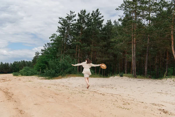 Back view of young woman with outstretched hands holding straw hat and walking near forest — Stock Photo