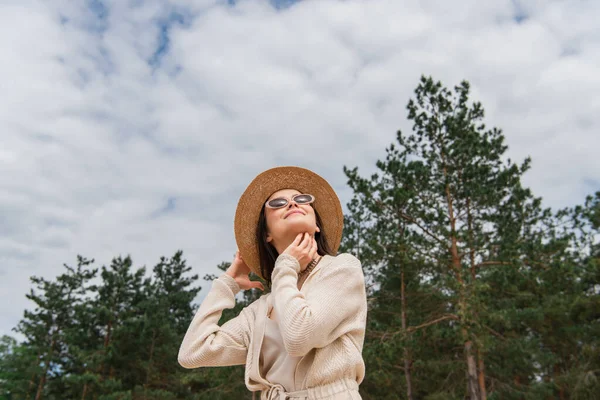 Visão de ângulo baixo da jovem mulher alegre em óculos de sol e chapéu de palha olhando para cima perto da floresta — Fotografia de Stock