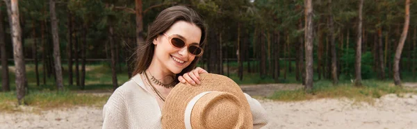 Alegre joven mujer en gafas de sol sosteniendo sombrero de paja cerca del bosque, bandera - foto de stock