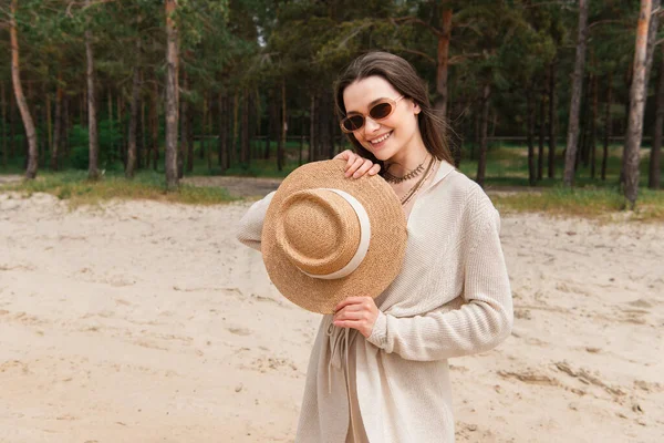 Cheerful young woman in sunglasses holding straw hat near green forest — Stock Photo