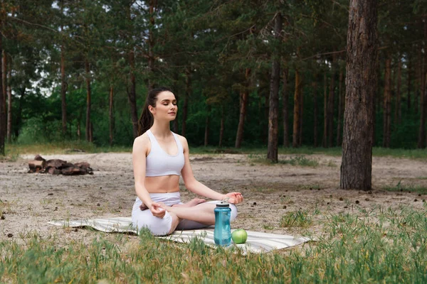 Paisible jeune femme en vêtements de sport assis dans la pose de lotus et méditant sur tapis de yoga — Photo de stock