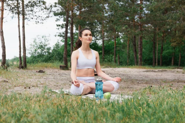 Joven mujer en ropa deportiva sentada en pose de loto y meditando en esterilla de yoga - foto de stock