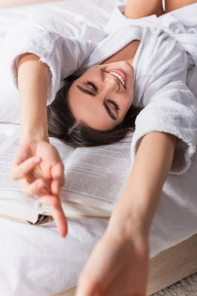 Fröhliche junge Frau mit verschwommenen ausgestreckten Händen neben Buch auf dem Bett liegend — Stockfoto