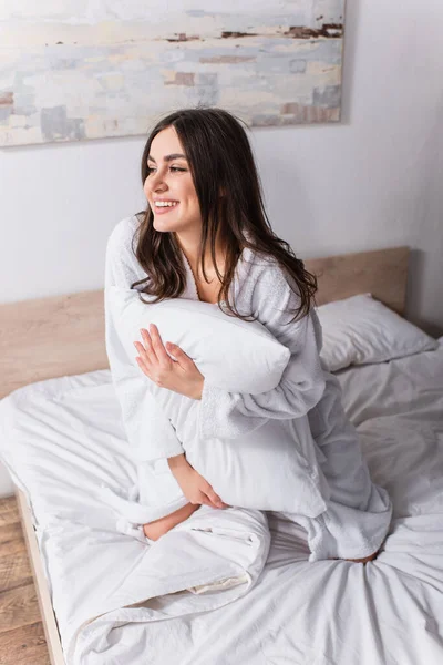 Задоволена жінка в халаті тримає подушку, сидячи на ліжку — стокове фото