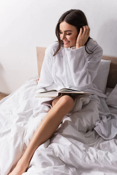 Mujer sonriente ajustando el cabello mientras lee novela en el dormitorio - foto de stock