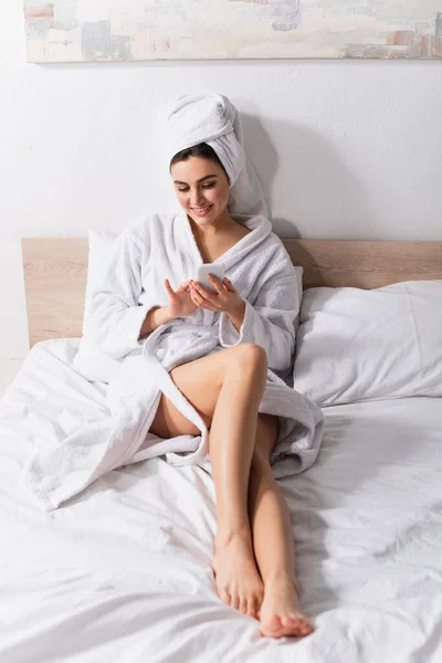 Happy woman in towel on head and bathrobe using smartphone in bedroom — Stock Photo