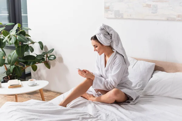 Mujer feliz en toalla en la cabeza y albornoz con teléfono inteligente en el dormitorio moderno - foto de stock