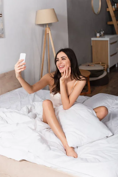 Alegre jovem mulher saindo da língua e tomando selfie enquanto sentado na cama — Fotografia de Stock