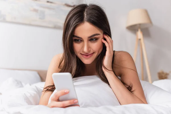 Joyful young woman lying on bed and using smartphone — Stock Photo