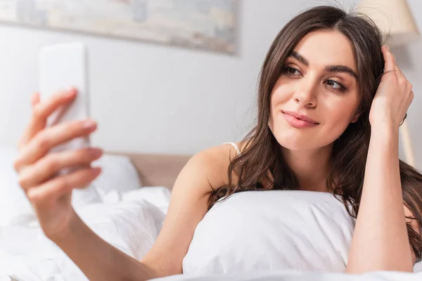 Joyeuse jeune femme couchée sur le lit et prenant selfie sur smartphone — Photo de stock