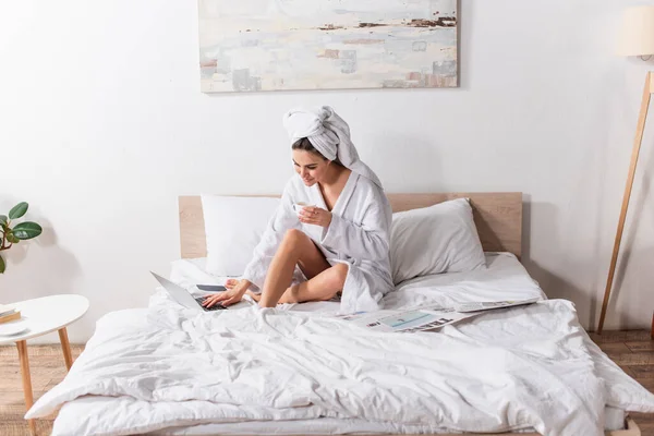 Happy woman in bathrobe and towel on head holding cup of coffee and using laptop near smartphone and newspaper on bed — Stock Photo