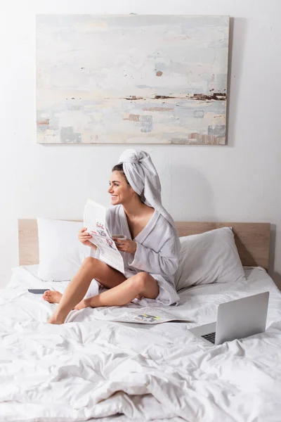 Mujer feliz en albornoz y toalla en la cabeza sosteniendo taza de café y periódico cerca de gadgets en la cama - foto de stock