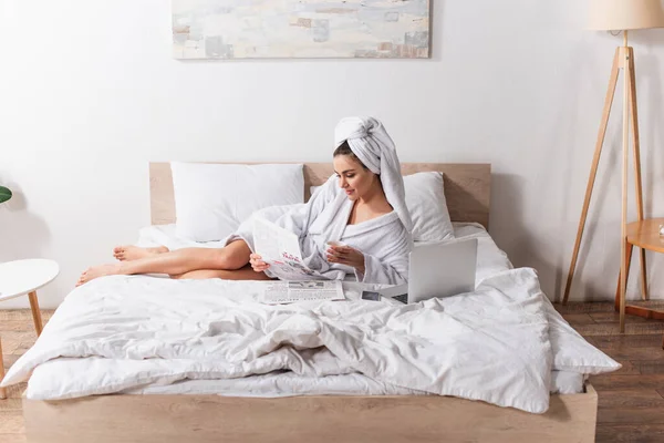 Mujer joven en albornoz y toalla en la cabeza sosteniendo taza de café y periódico cerca de gadgets en la cama - foto de stock