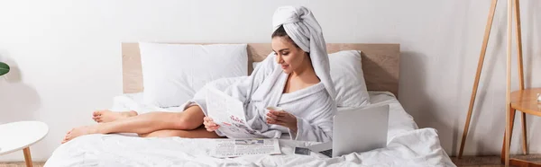 Молодая женщина в халате и полотенце на голове держа чашку кофе и газету возле гаджетов на кровати, баннер — стоковое фото