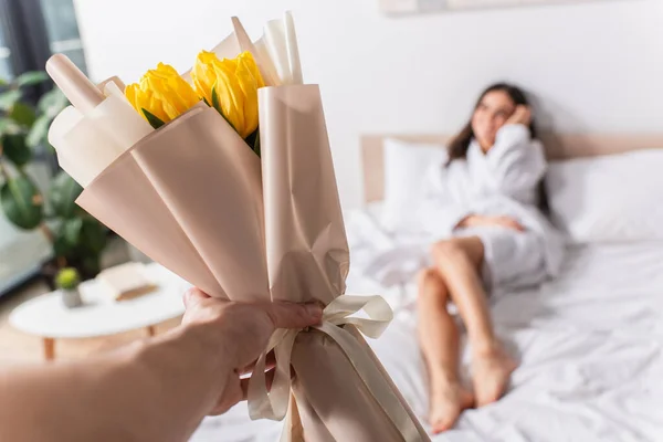 Mann hält Strauß gelber Tulpen neben verschwommener Frau im Schlafzimmer — Stockfoto